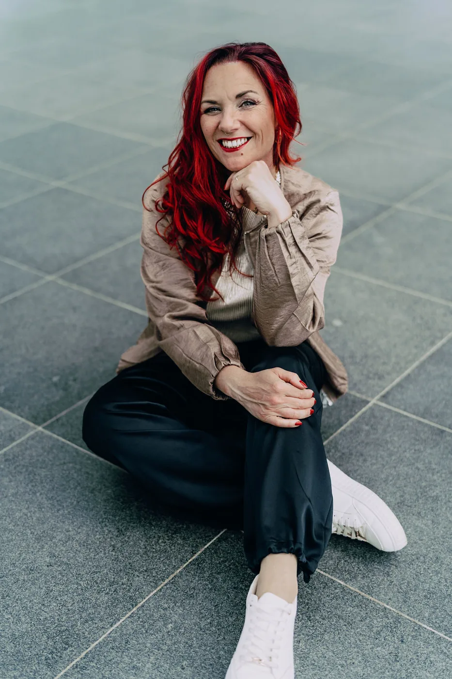Sabrina Amels sitzt lachend auf dem Steinboden, das Kinn auf die Faust gestützt.